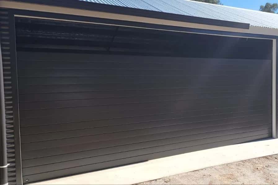 A closed dark grey sectional garage door on a ribbed steel-clad shed under a clear sky