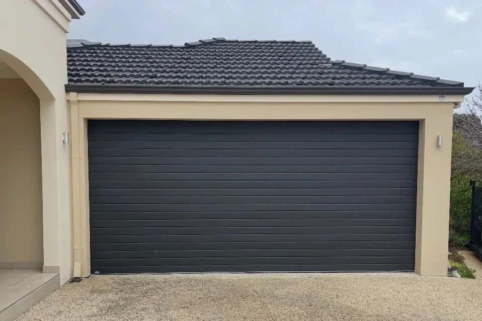 A closed charcoal double garage door with narrow horizontal ribs, above an exposed-aggregate driveway