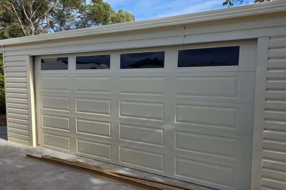 A cream sectional garage door with a row of tinted windows, closed in a matching clad shed