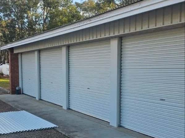 Four closed white roller doors in a row along a long shed, with gum trees and cloud behind