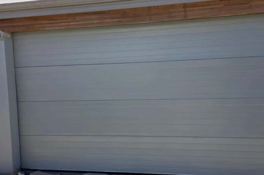 A pale grey sectional garage door closed under a raw timber fascia beam