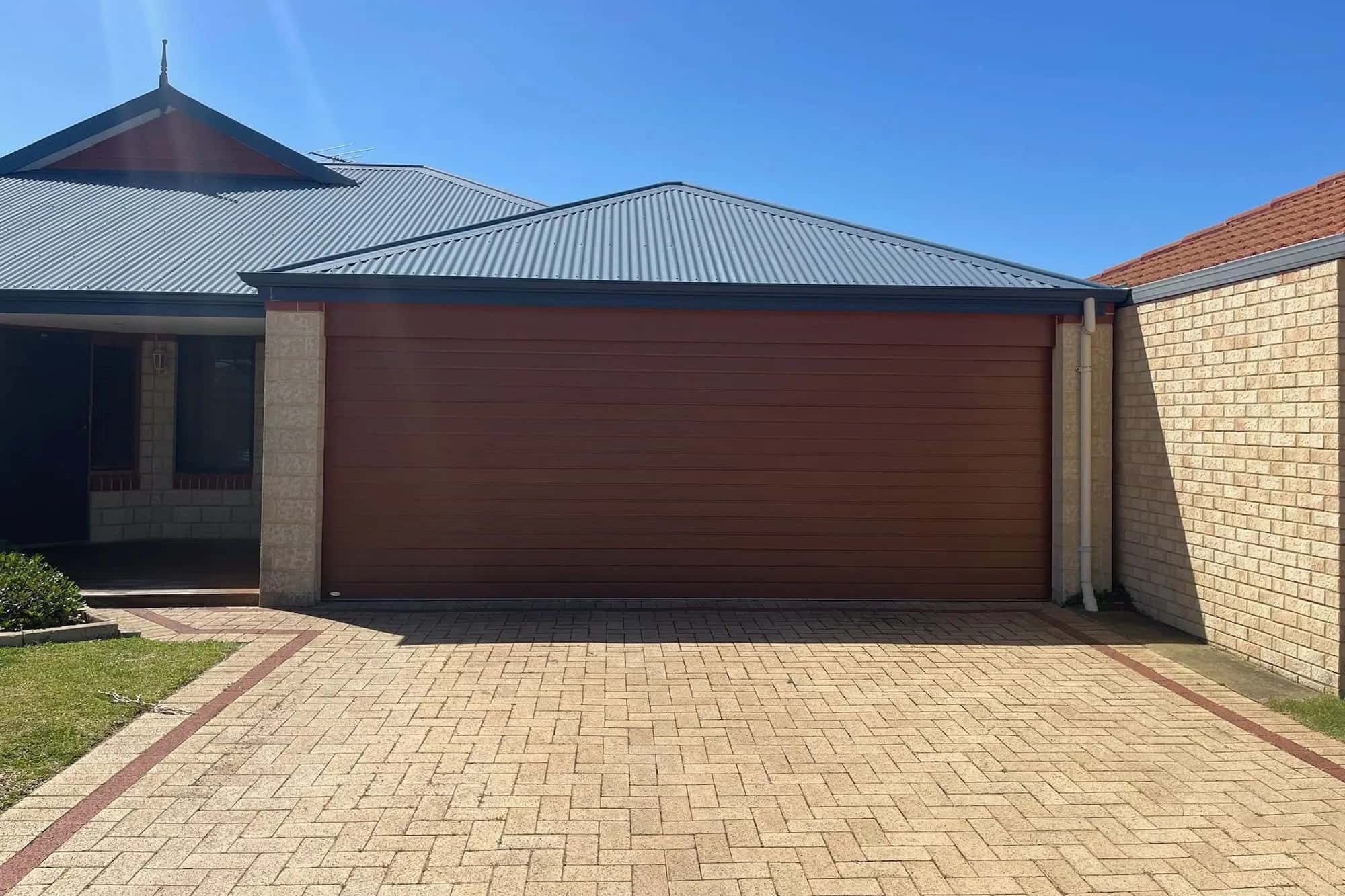 A new red-brown flat-panel double sectional garage door between cream brick piers, with a herringbone paved driveway