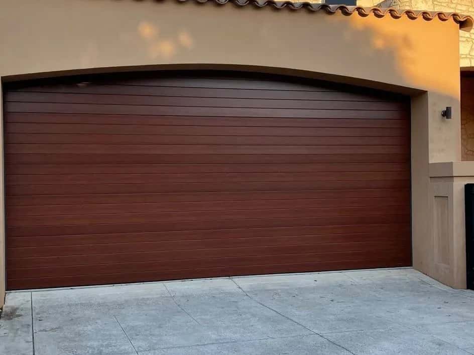 A woodgrain-look double sectional garage door fitted to an arched opening in a rendered wall, in late afternoon sun