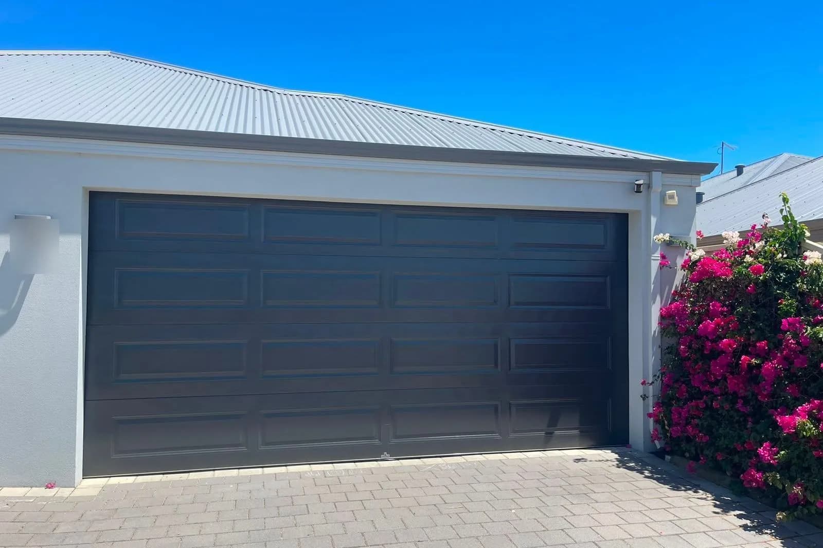 A charcoal raised-panel sectional door under a white roof, with bougainvillea in flower beside it