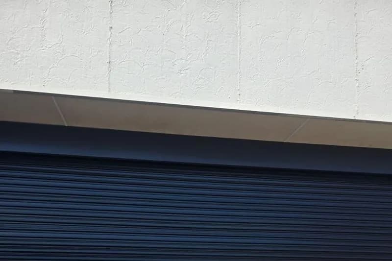Close-up of a closed charcoal roller door curtain, its horizontal slats running under a white rendered wall and a pale concrete lintel