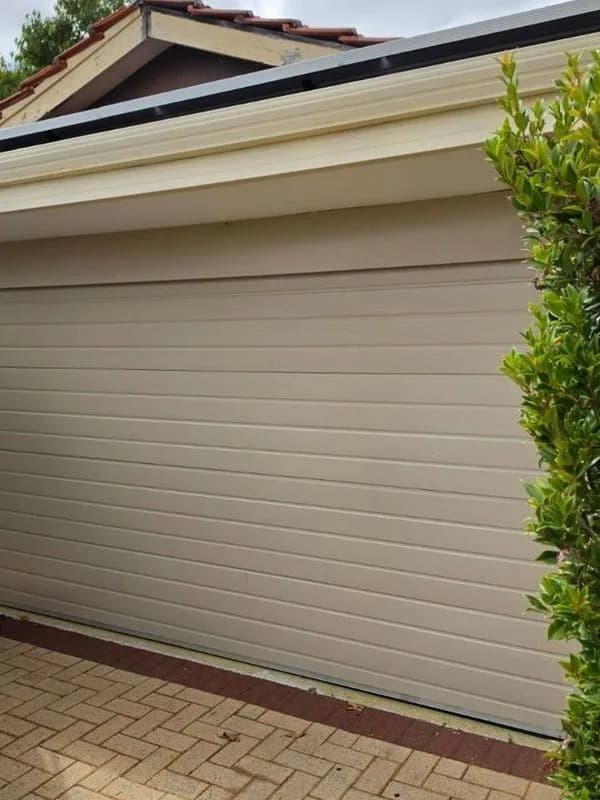 A closed cream roller door on a single garage, seen at an angle beneath a tiled gable, with herringbone brick paving in front