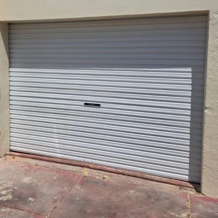 A closed white roller garage door set into a cream rendered wall, with red brick paving in front