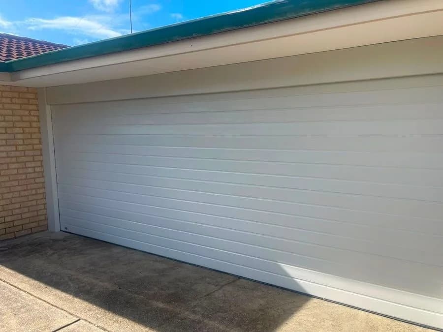 A white ribbed sectional garage door closed onto a concrete driveway in bright sun, a brick pier on the left of the opening