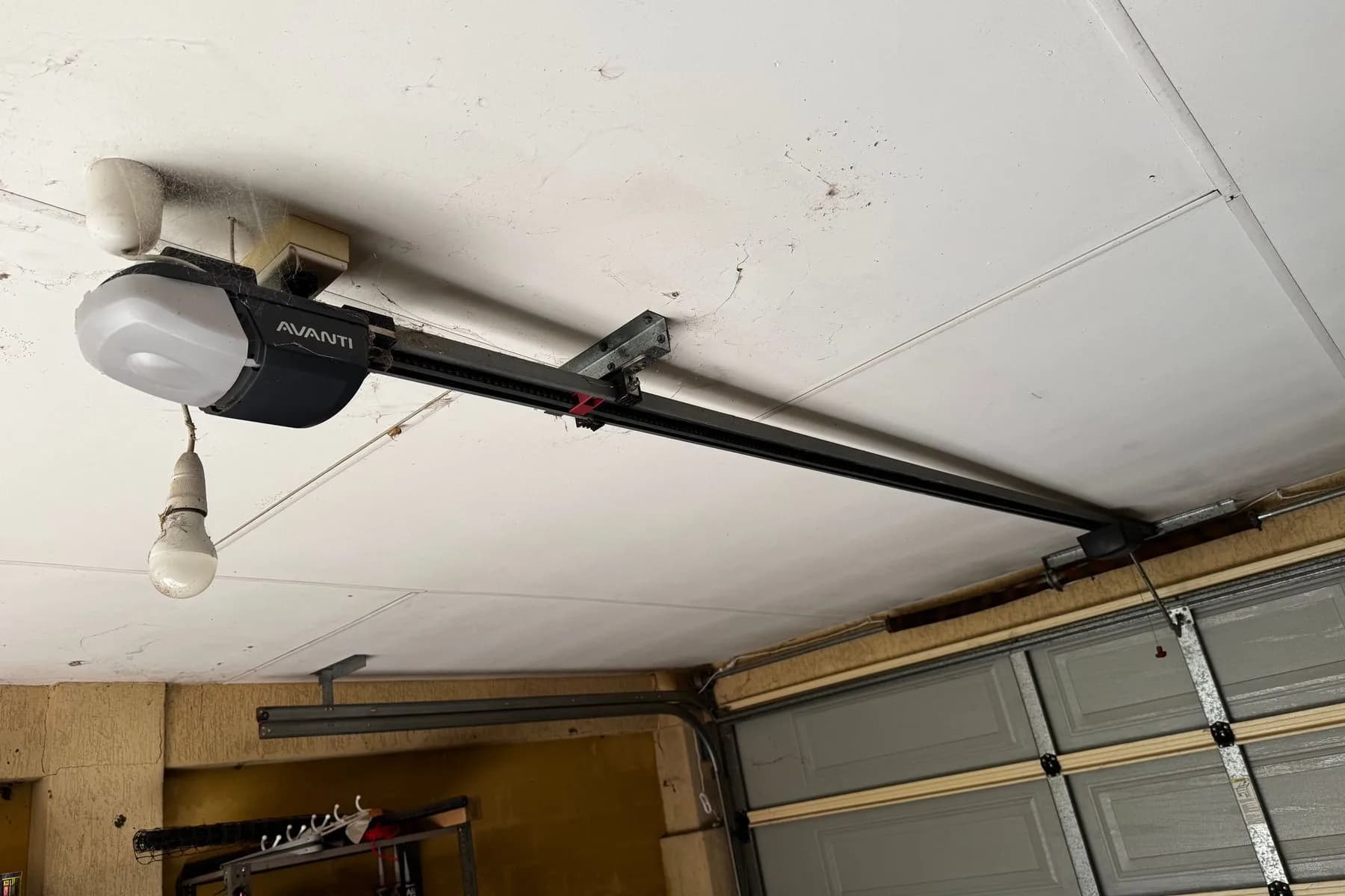 A garage door opener rail running from the motor unit to the top of a grey sectional door, the red manual-release handle hanging near the door
