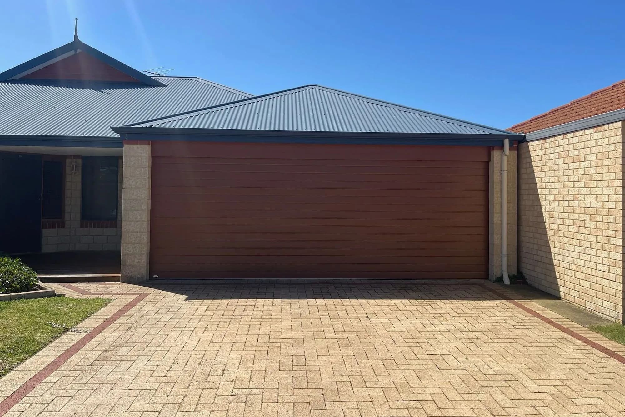 A new red-brown flat-panel double sectional garage door between cream brick piers, with a herringbone paved driveway