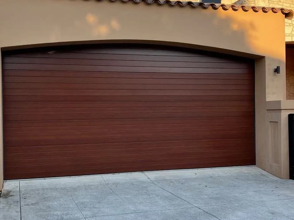A woodgrain-look double sectional garage door fitted to an arched opening in a rendered wall, in late afternoon sun