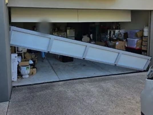 A white sectional garage door hanging diagonally across the opening after coming out of its tracks, one end resting on the garage floor with boxes and shelving visible behind it
