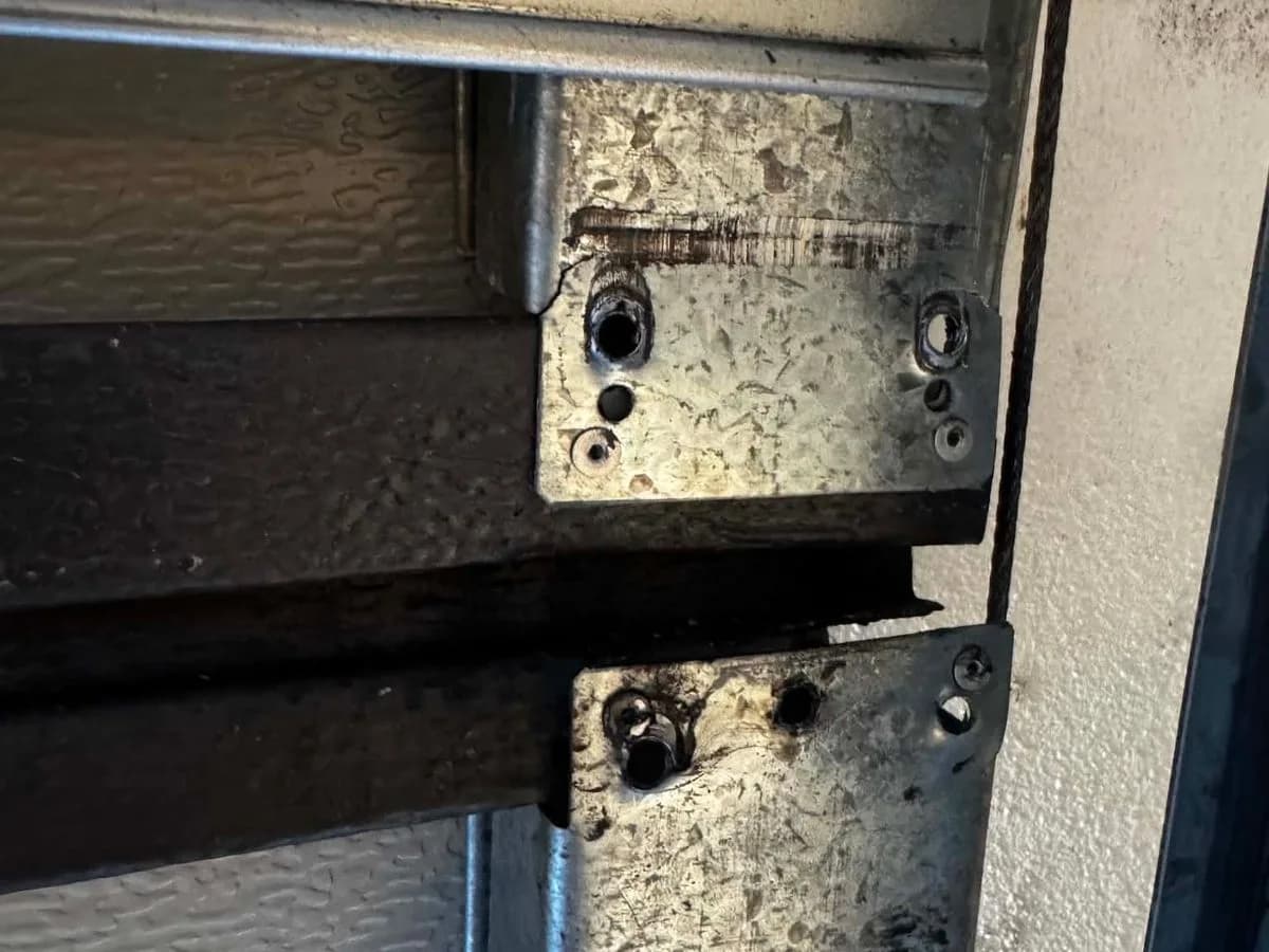 Two worn galvanised hinge plates on a sectional garage door panel, the upper one with a fixing hole wallowed out and the metal scored where the roller stem has been working loose