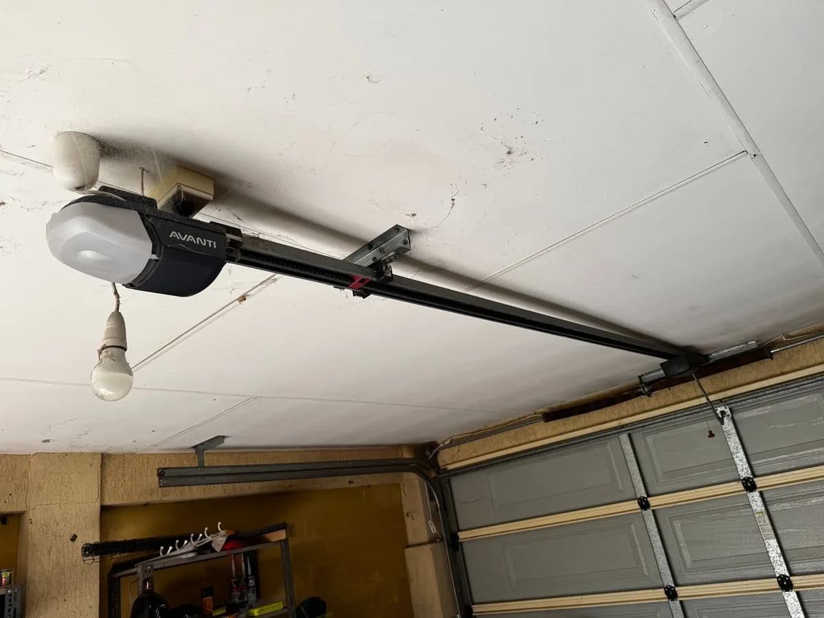 An older ceiling-mounted garage door opener on its rail above a grey sectional door, a bare bulb hanging beside the motor unit