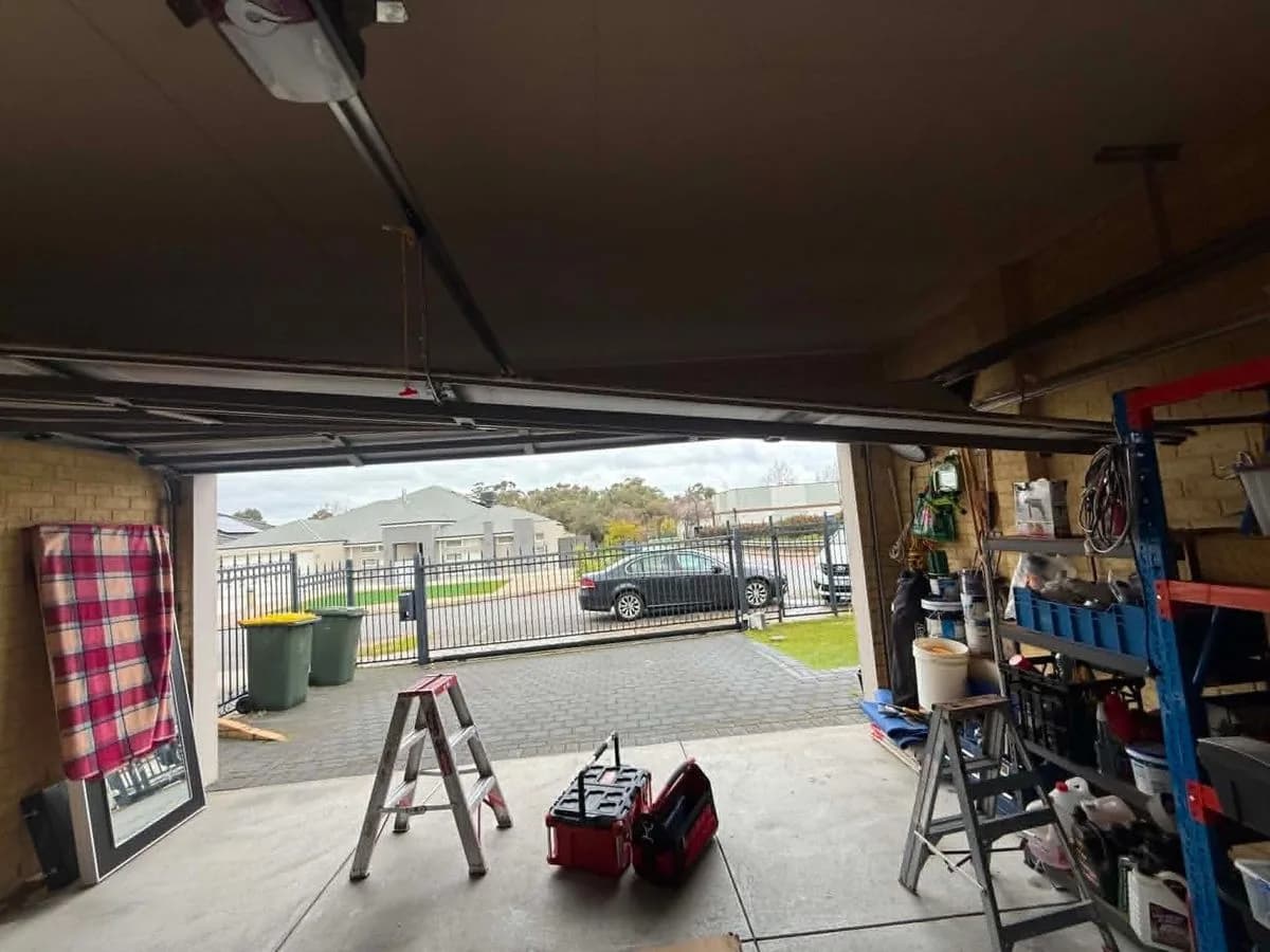 Looking up from inside a garage at the opener rail and motor mounted under the ceiling, with the sectional door raised and a stepladder and toolbox on the floor below