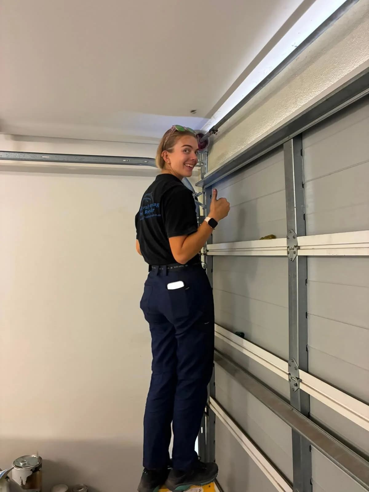 A technician on a ladder beside a sectional door track, turning to give a thumbs up