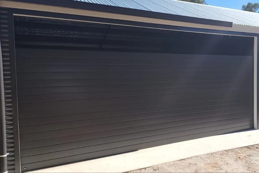 A closed dark grey sectional garage door on a ribbed steel-clad shed under a clear sky A closed dark grey sectional garage door on a ribbed steel-clad shed under a clear sky
