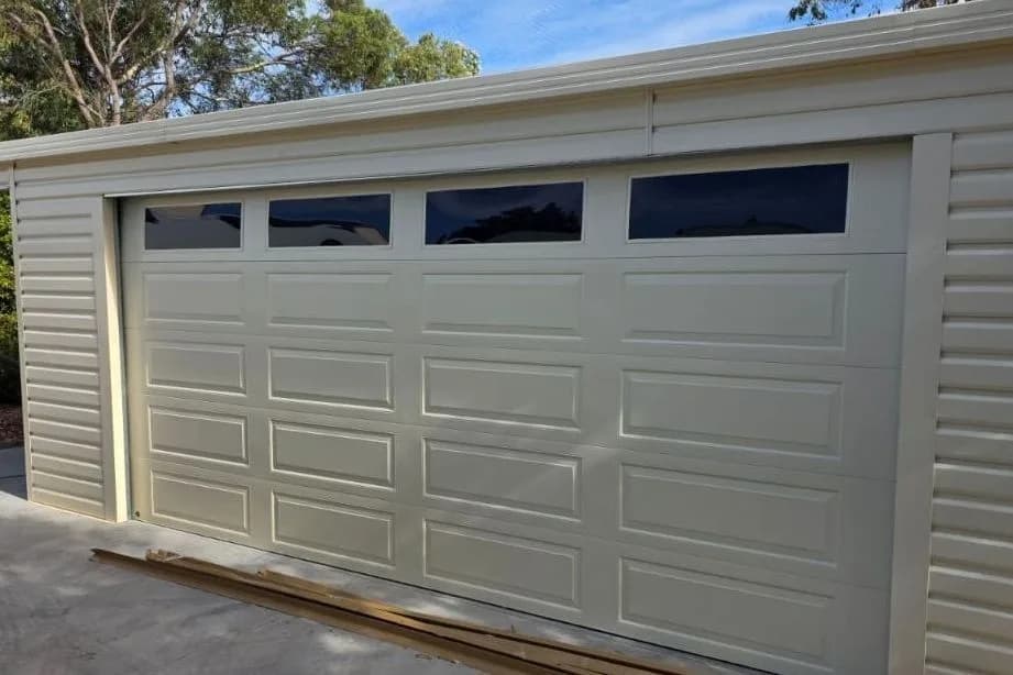 A cream sectional garage door with a row of tinted windows, closed in a matching clad shed A cream sectional garage door with a row of tinted windows, closed in a matching clad shed