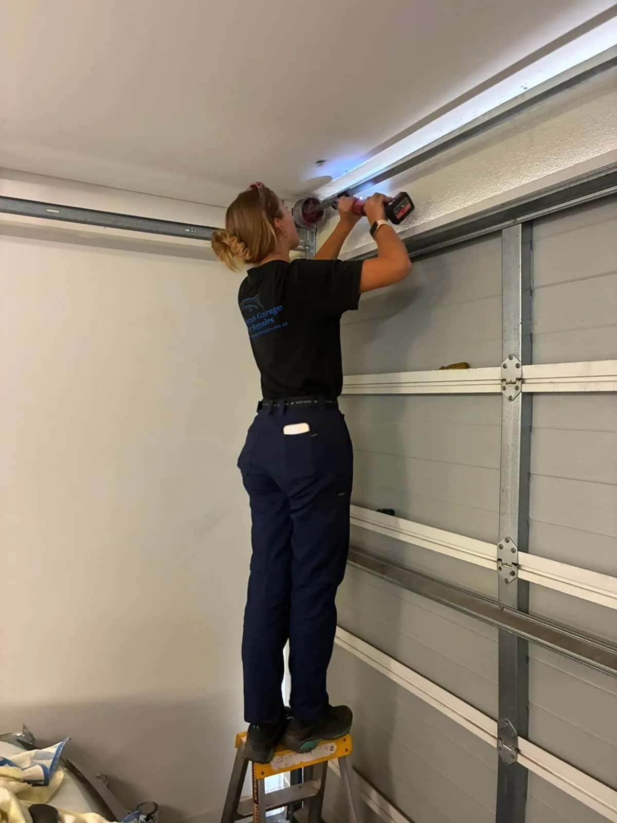 A technician on a step ladder drilling into the top track of a sectional garage door