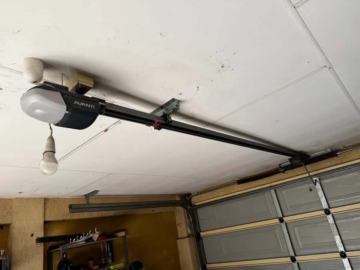 An old ceiling-mounted opener on its rail in a cluttered garage, a bare bulb hanging beside it